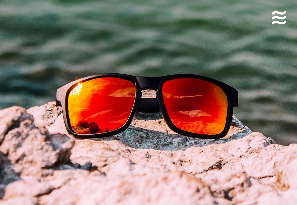 Front view of black sunglasses with red mirrored lenses showcasing rectangular frame on sunlit rock by green water