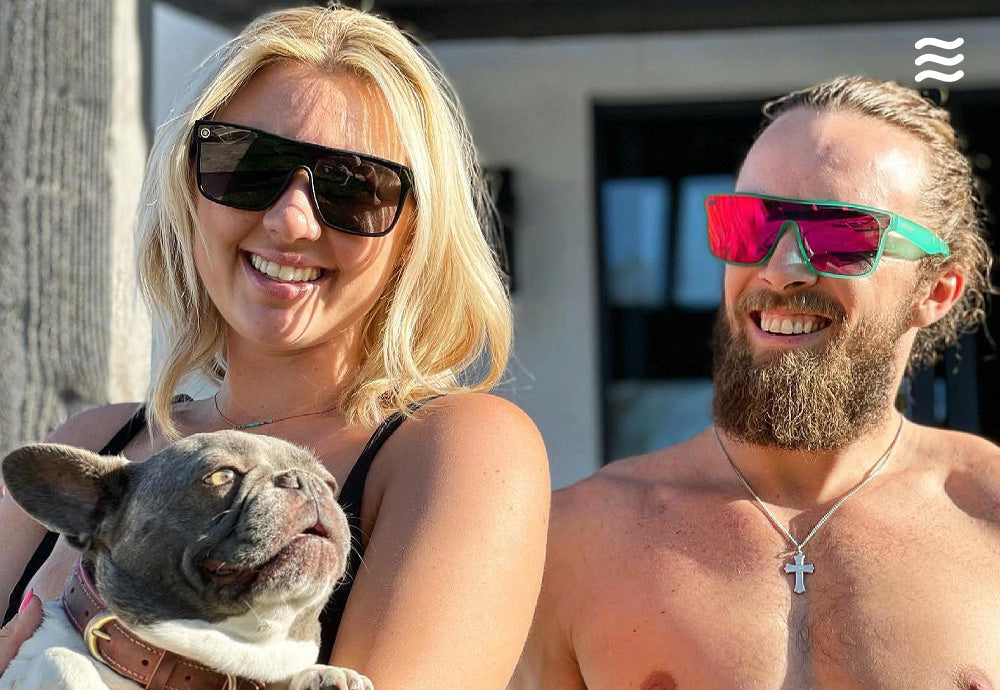 Smiling woman and bearded man wearing sunglasses pose outdoors with a French bulldog on the woman’s lap