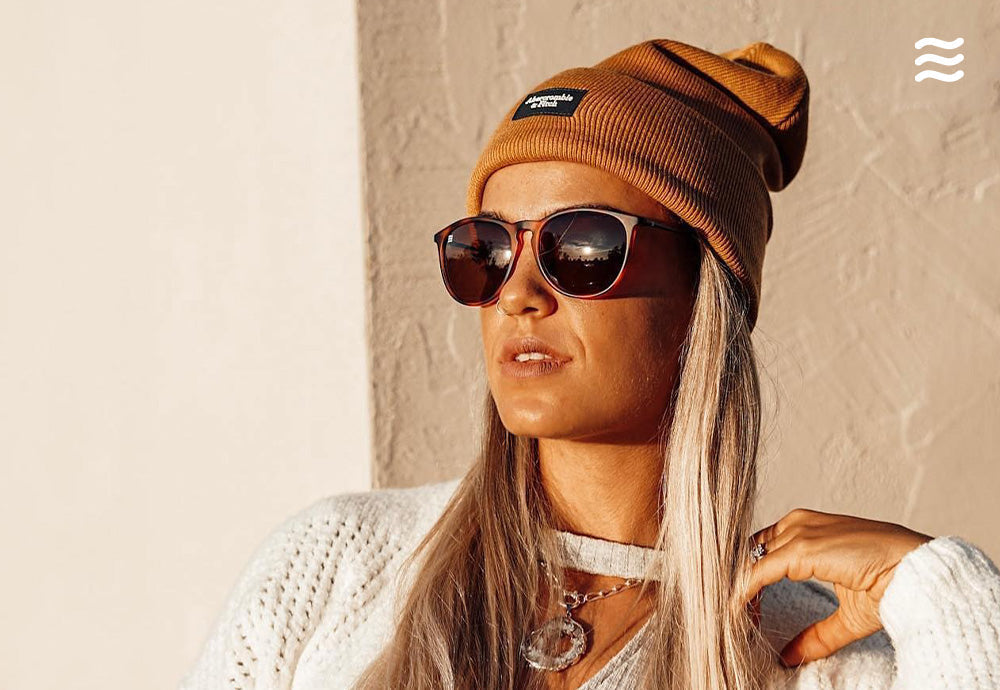Woman wearing brown sunglasses and a mustard beanie posing against a sunlit textured wall