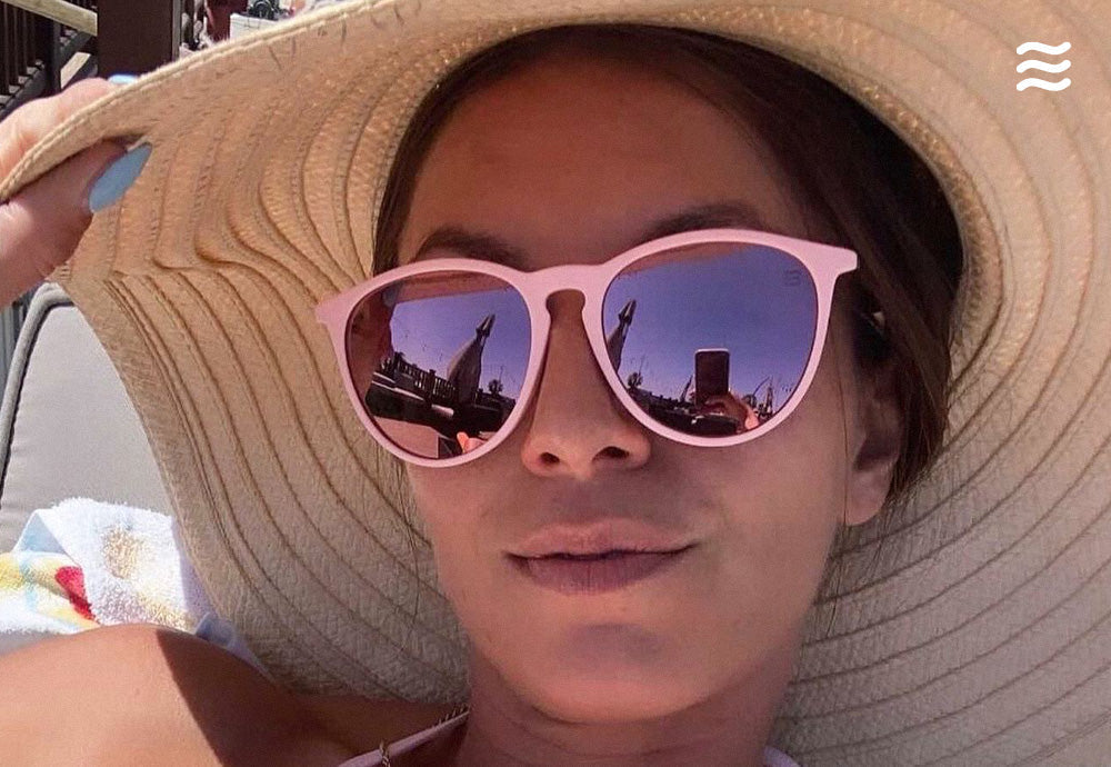 Woman reclining on a lounge chair wearing pink-framed mirrored sunglasses and a wide-brim straw sun hat with a towel visible
