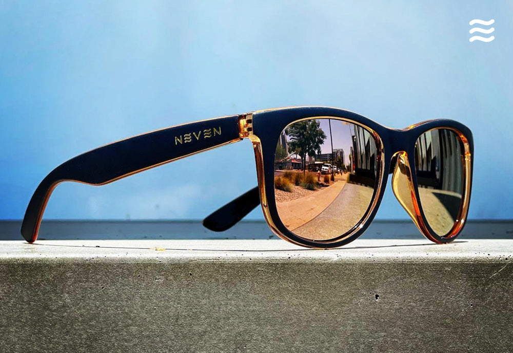 Side view of Neven mirrored sunglasses with gold lenses and black-and-amber frame on concrete ledge against blue sky