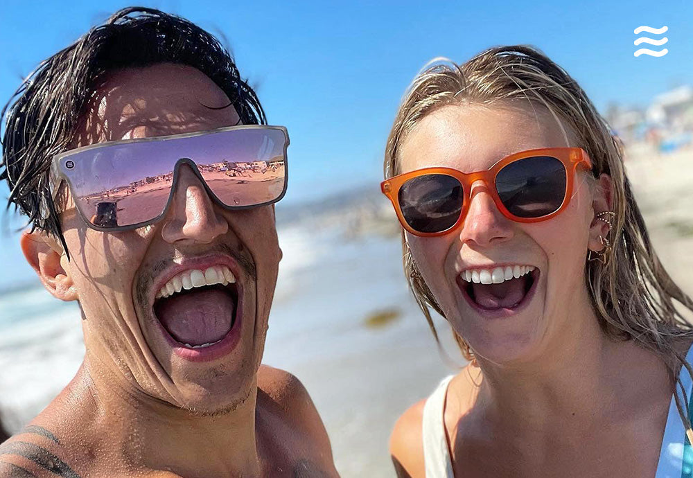 Two people laughing on a sunny beach wearing a mirrored shield sunglass and orange round sunglasses