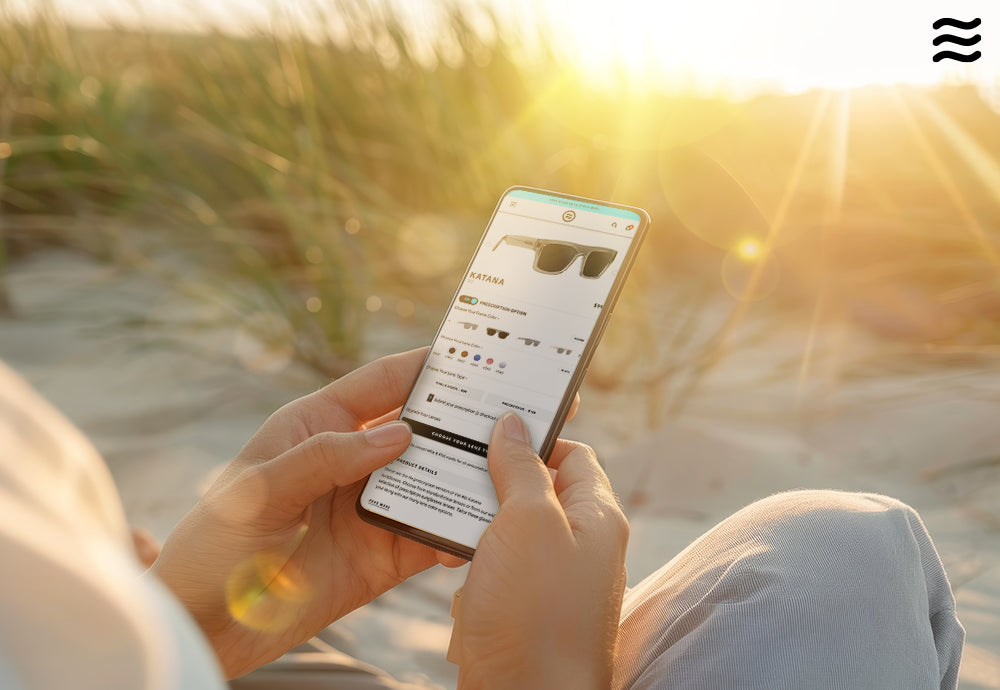 Person holding smartphone browsing a prescription sunglasses product page on a sunlit beach at sunset