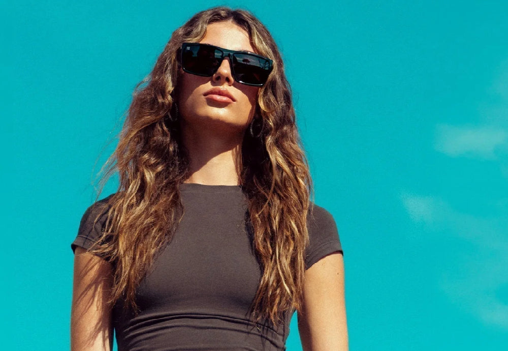 Woman wearing oversized black rectangular sunglasses and a fitted charcoal t-shirt standing against a clear blue sky