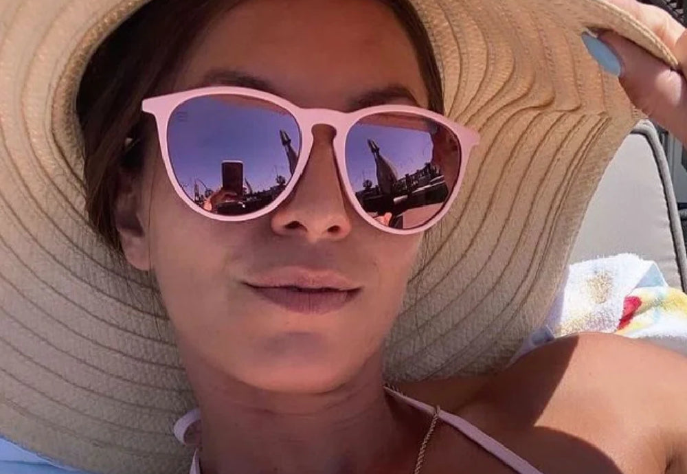 Woman sunbathing under a wide-brim straw hat wearing pink-framed mirrored sunglasses