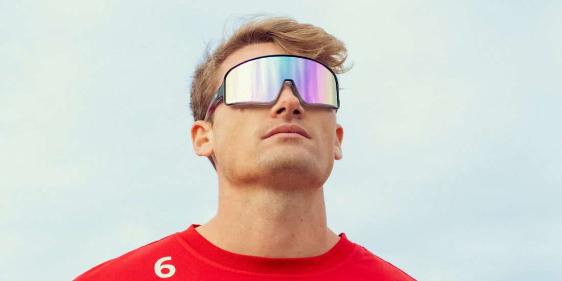 Man wearing large black and purple G.O.A.T. polarized sunglasses outdoors against a pale blue sky