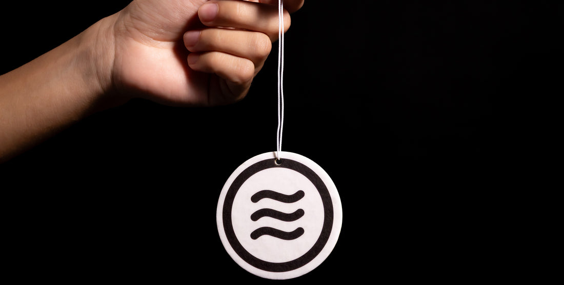 View of Signature Air Freshener showing Neven tilde logo, 2.5-inch disc hanging from string held by hand on black background