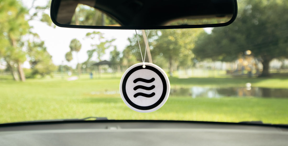 Front of Signature Air Freshener showing black Neven tilde logo on 2.5 inch disc hanging from car mirror, blurred park background