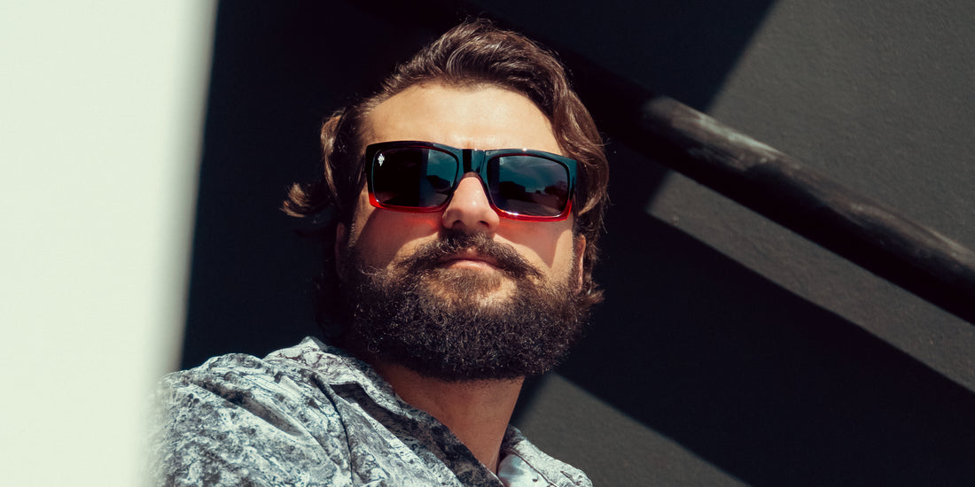 Bearded man wearing Blood Fade polarized sunglasses with black-to-red fade, looking down from a sunlit stairwell