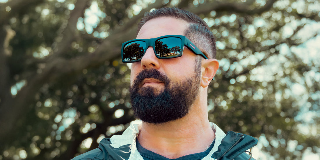 Bearded man wearing Redrum polarized black rectangular sunglasses looking up with trees reflected in the lenses