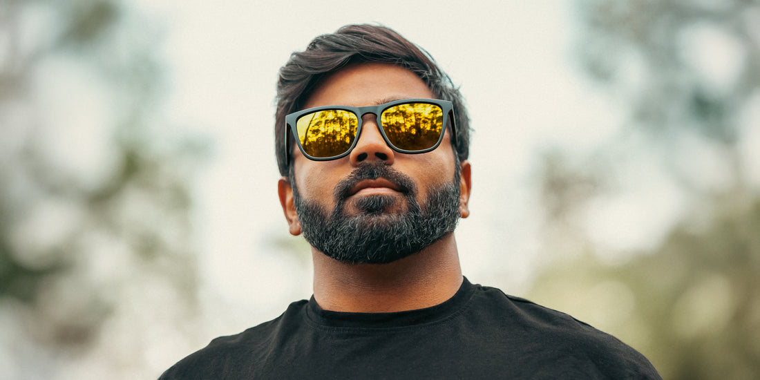Bearded man wearing Liquid Gold black sunglasses outdoors with blurred trees in the background
