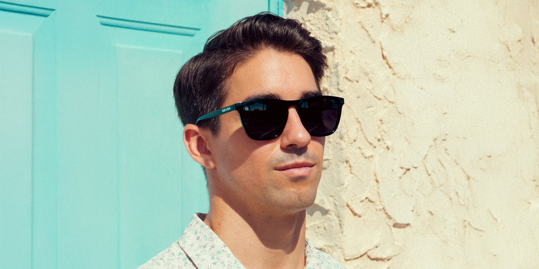 Man wearing Nix black polarized rectangle sunglasses standing by a turquoise door and sunlit stucco wall