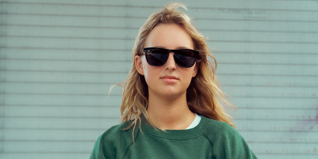 Woman wearing Nix medium black rectangular polarized sunglasses against a pale blue horizontal-paneled wall