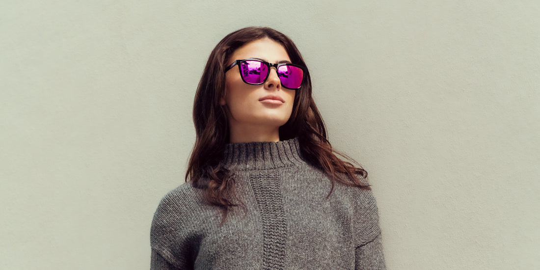 Woman wearing black-framed Cambridge rectangular sunglasses with deep purple mirrored lenses, looking up against a pale textured wall