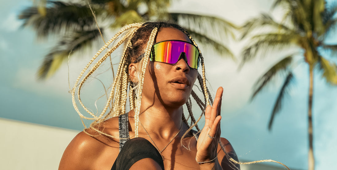 Woman posing outdoors in FLL SND wraparound sunglasses with pink to yellow gradient lenses and palm trees in the background