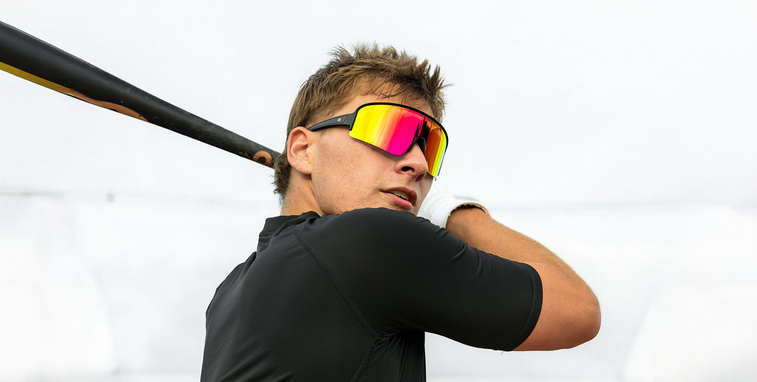 Man wearing FLL SND wraparound sunglasses with pink to yellow gradient lenses, turning outdoors in a black shirt