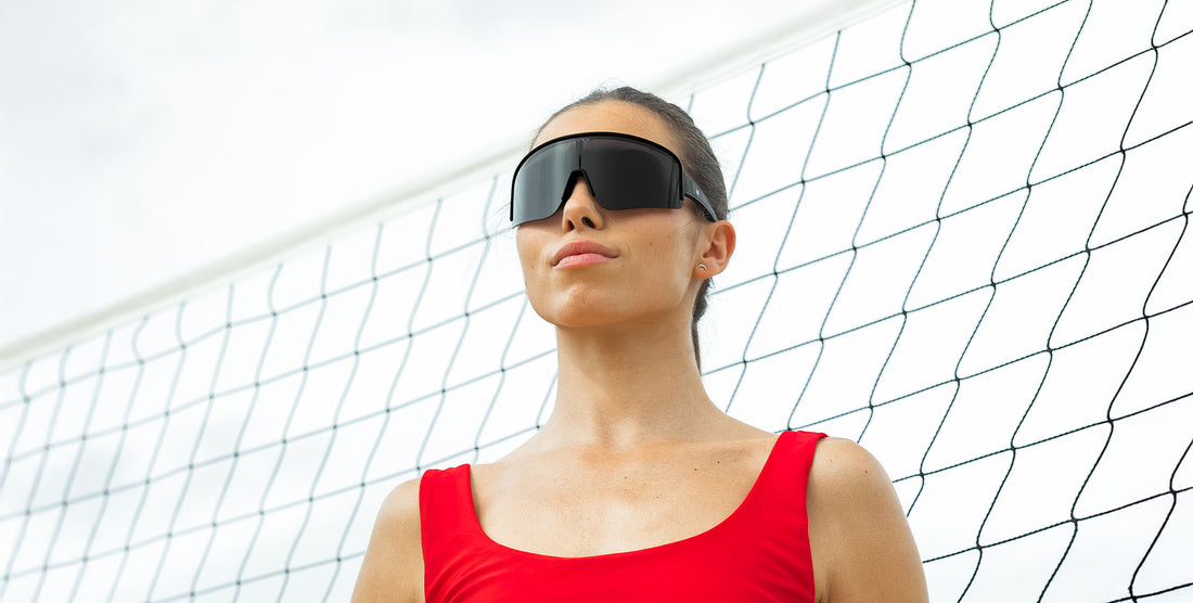 Model wearing Threshold black polarized wraparound sunglasses with straight arms, standing in front of a sports net