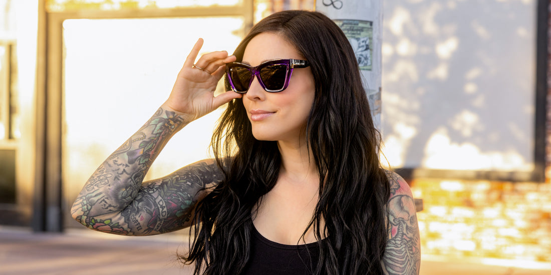Tattooed woman adjusting Ethereal purple cat-eye sunglasses with glossy frame and black polarized lenses at golden hour