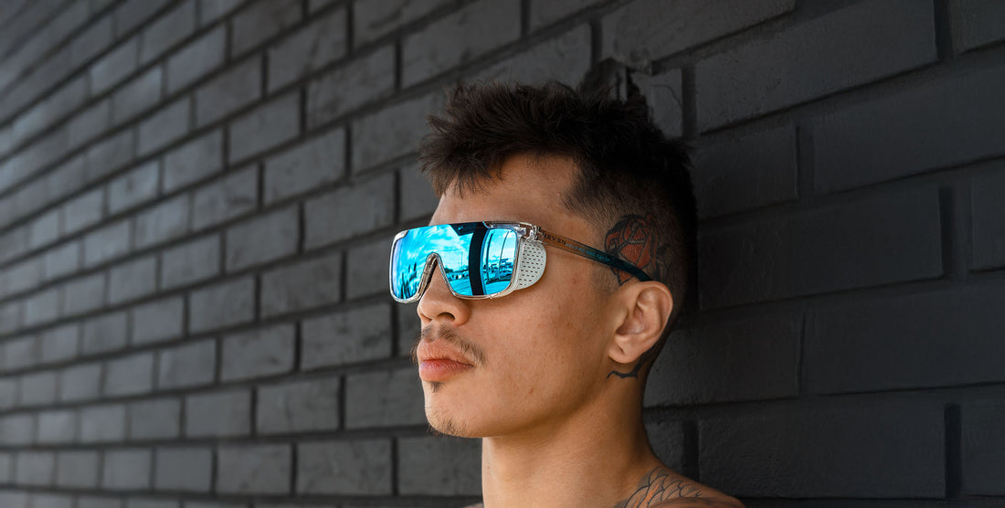 Man wearing Iced Earth clear rectangular sunglasses with mirrored blue polarized lenses against a dark brick wall