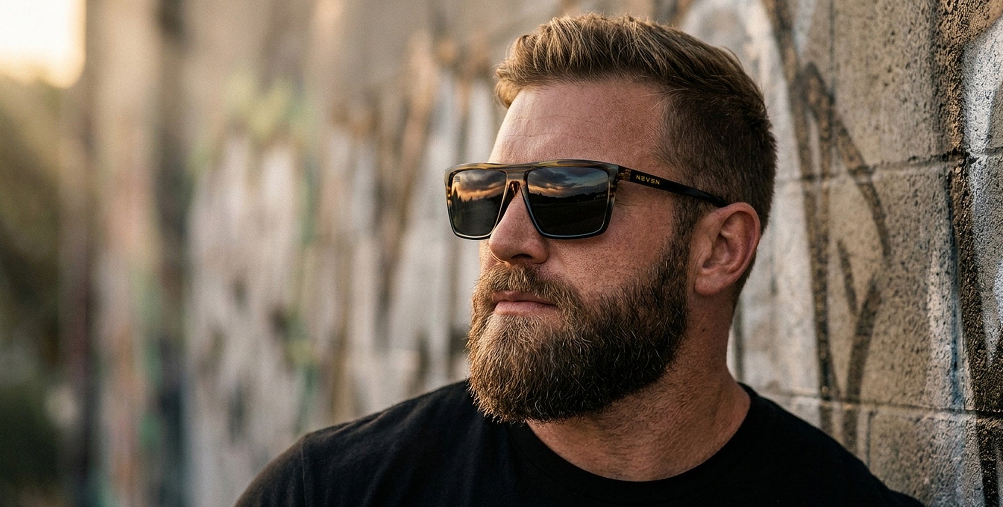 Man wearing Asbury XL black wood-grain polarized sunglasses leaning against a graffiti-covered wall at sunset
