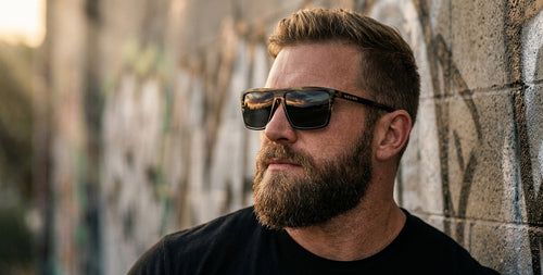 Man wearing Asbury XL black wood-grain polarized sunglasses leaning against a graffiti-covered wall at sunset