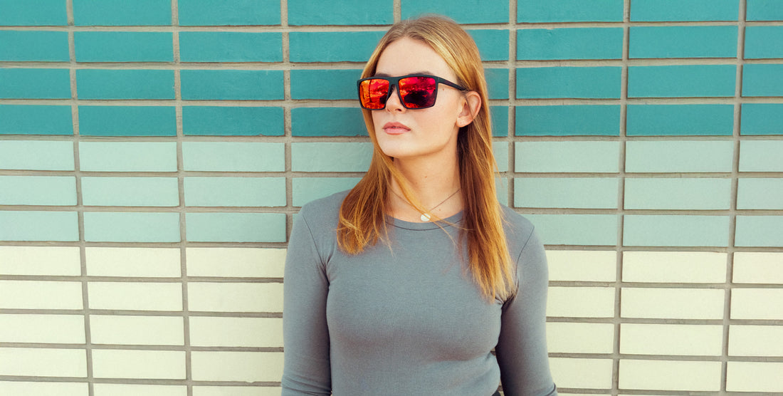 Woman wearing 212 sunglasses with black square frame and red polarized mirrored lenses, standing against a teal-tiled wall