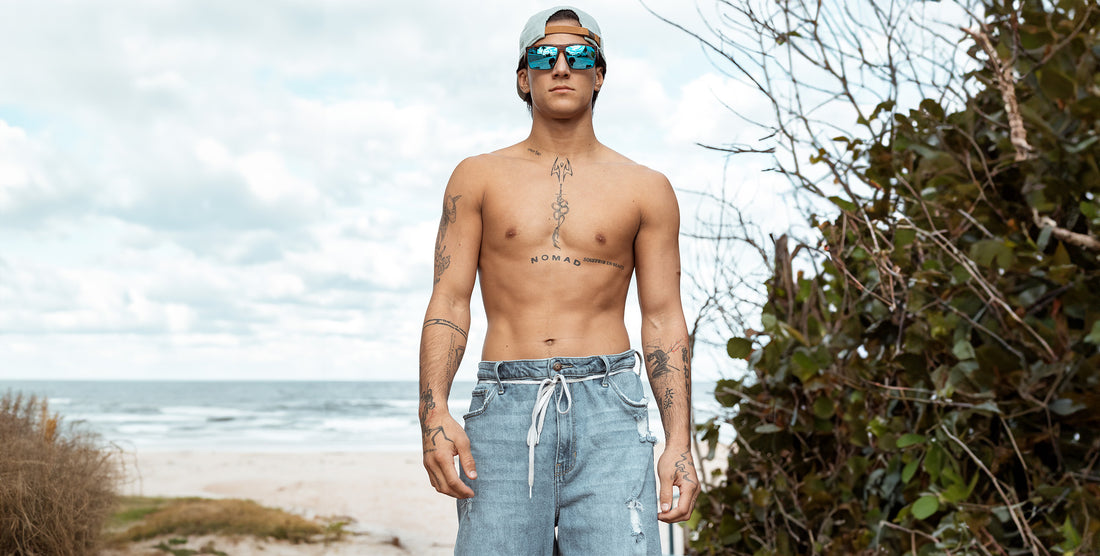 Shirtless man standing on a beach wearing Melby polarized sunglasses with blue mirrored lenses and wood-patterned frame