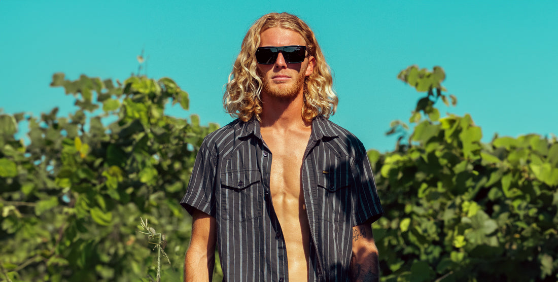 Male model wearing NYC black rectangular polarized sunglasses standing in front of leafy greenery under a bright blue sky