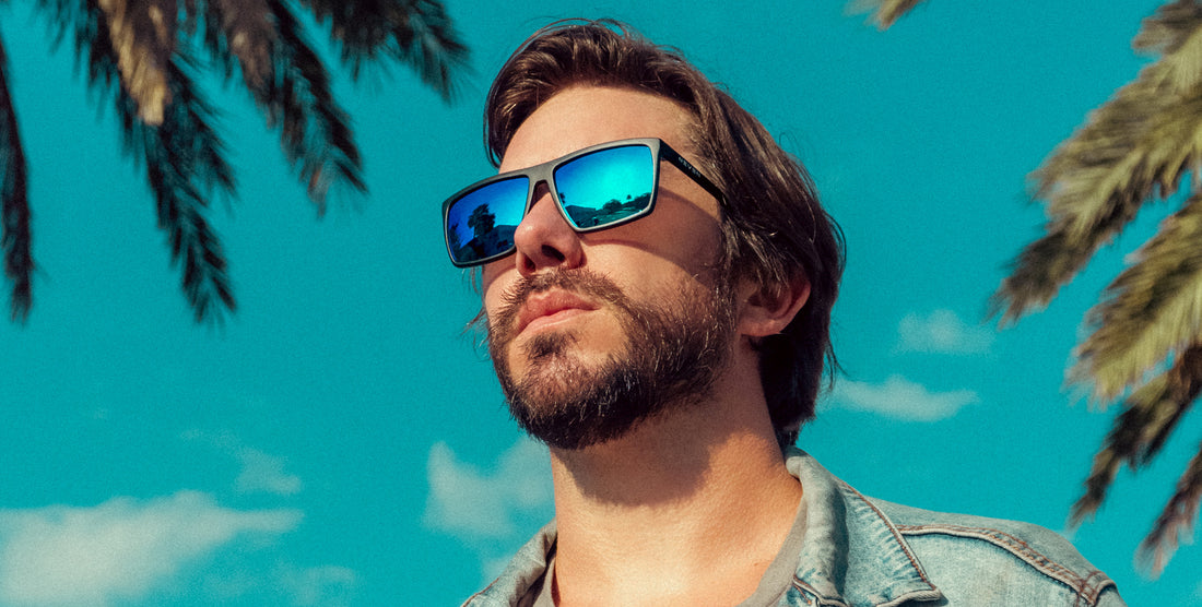 Man wearing Three Of Cups black polarized sunglasses with blue-green lenses, looking up against a sunny sky with palm trees