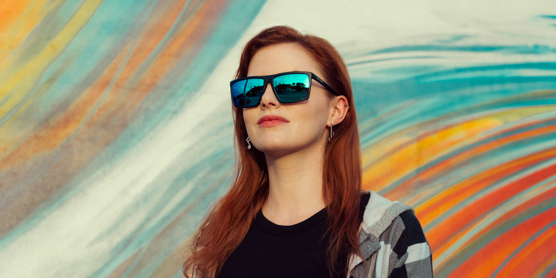 Woman wearing Three Of Cups polarized sunglasses with matte black frames and blue-green mirrored lenses in front of a colorful striped mural
