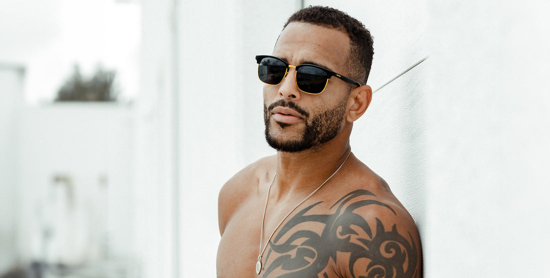 Man wearing Gold Rush black polarized sunglasses with gold trim leaning shirtless against a white wall in a bright outdoor setting