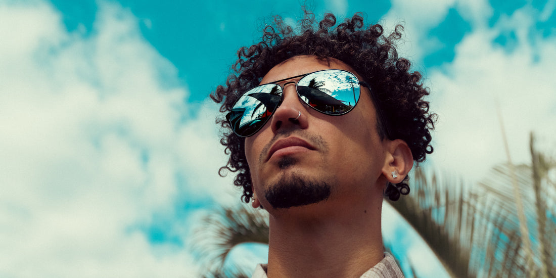 Man wearing black Mavs polarized sunglasses outdoors beneath a blue sky and palm trees