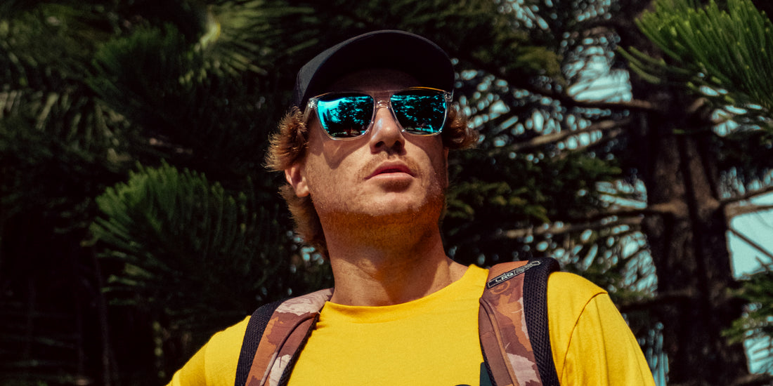 Man wearing clear-frame Lucid sunglasses with blue mirrored lenses looking up against sunlit pine trees, wearing a yellow shirt and backpack