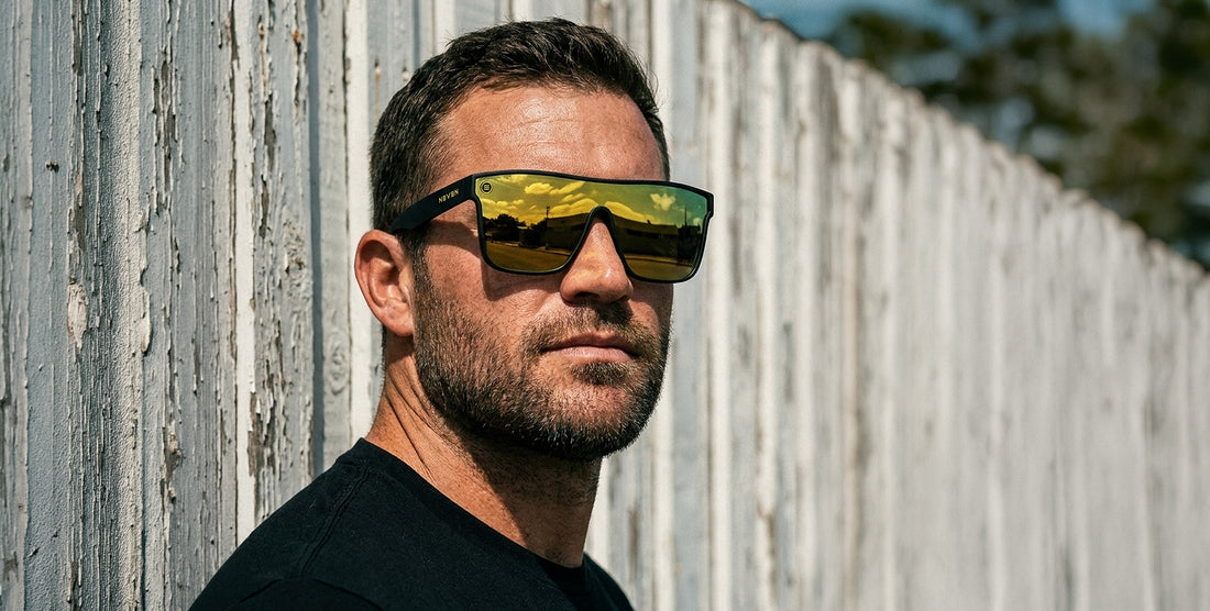 Three-quarter view of CWM XL sunglasses on model showing oversized black frame and mirrored gold lenses with Neven logo against weathered fence