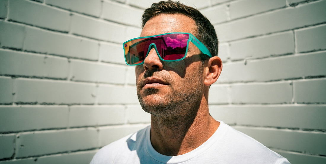 Close-up of X XL sunglasses with transparent teal frame and pink mirrored polarized lenses, worn against white brick wall