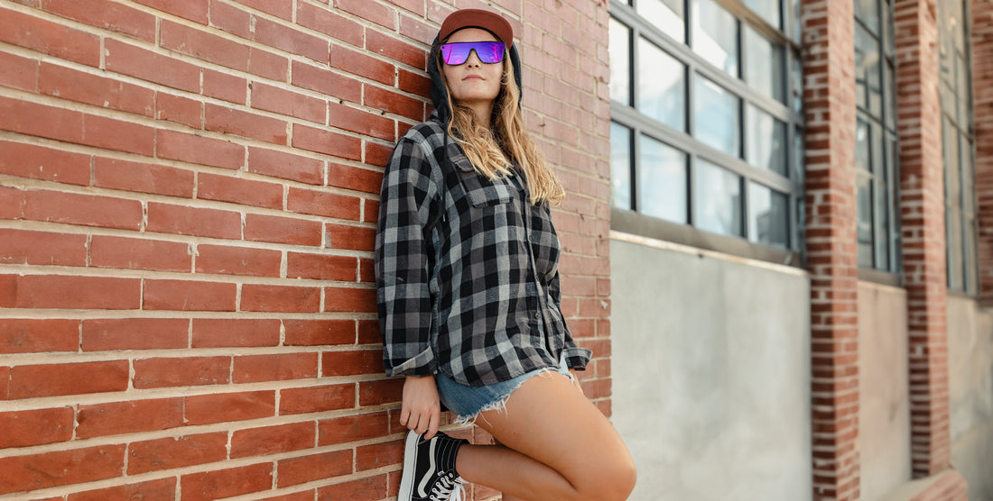 Woman wearing 53X gray square polarized sunglasses with purple mirrored lenses, leaning against a brick wall in a black-and-gray plaid shirt