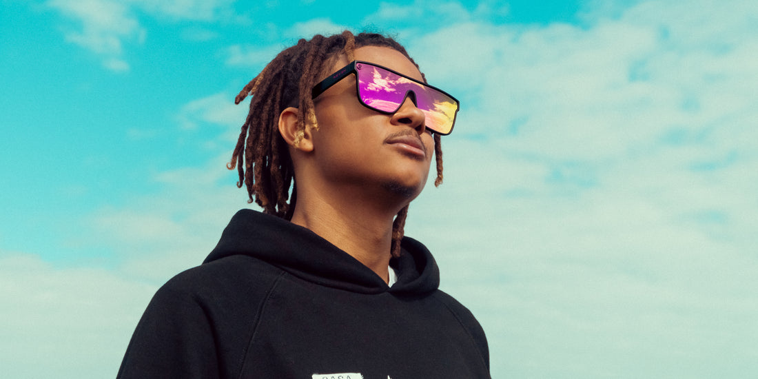 Model wearing Bliss sunglasses with black frame and pink/orange polarized lenses looking up against a bright blue sky