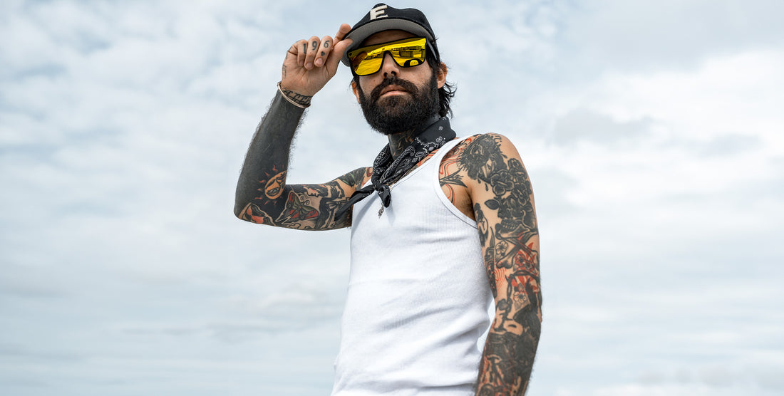 Tattooed man wearing CWM black-frame sunglasses with yellow mirrored unilens, tipping his cap against a cloudy sky