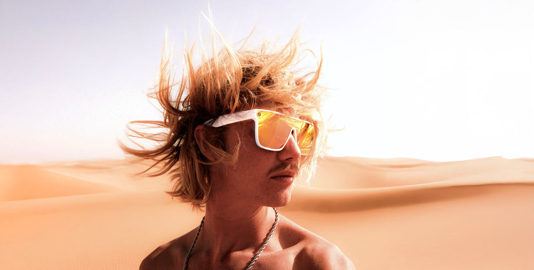 Man with windblown hair wearing white Muse oval polarized sunglasses with pink-yellow mirrored lenses in sunlit desert dunes