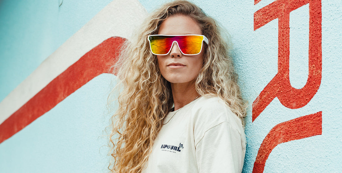 Woman wearing white Muse polarized sunglasses with pink and yellow lenses against a colorful painted wall