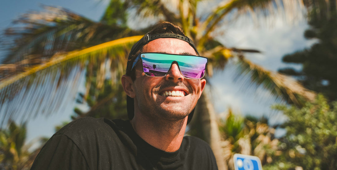 Man smiling while wearing Nomads Black Wayfarer polarized sunglasses outdoors with palm trees