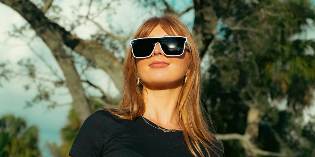 Woman wearing white Permafrost XL polarized sunglasses, looking up outdoors with trees reflected in the lenses