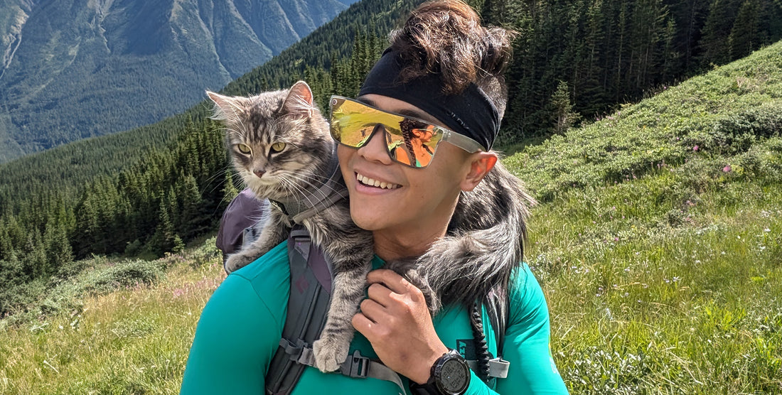 Person smiling on a mountain hike wearing Smooth Crush sunglasses with transparent gray frame and reflective pink/champagne polarized lens, a cat perched on their shoulders