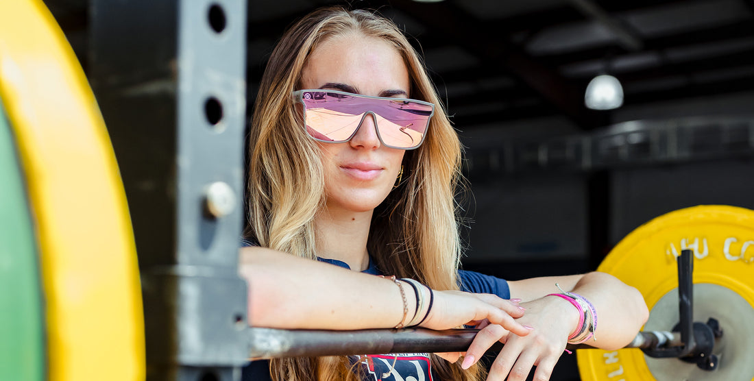 Model wearing Smooth Crush sunglasses leaning on a weight rack, showing transparent gray frame and pink mirrored polarized lens
