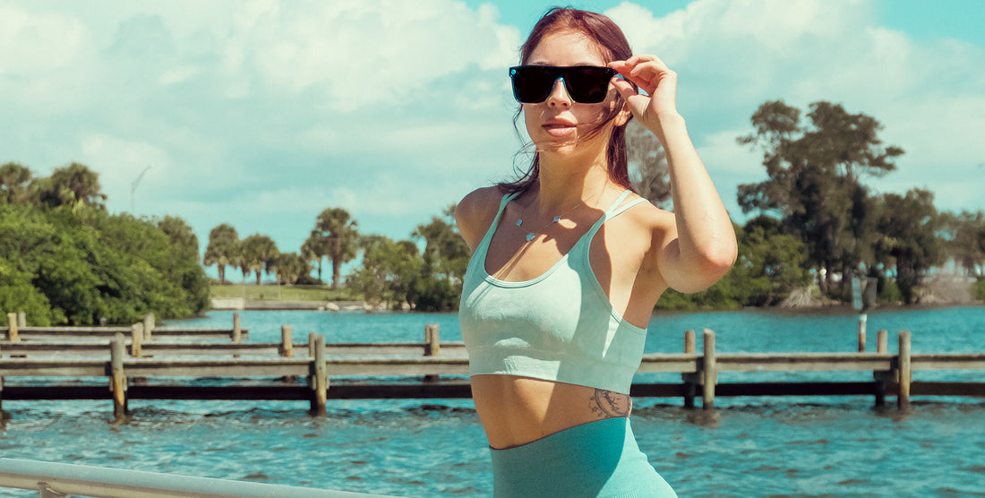 Woman adjusting black square sunglasses at a sunny dock with water and trees in the background