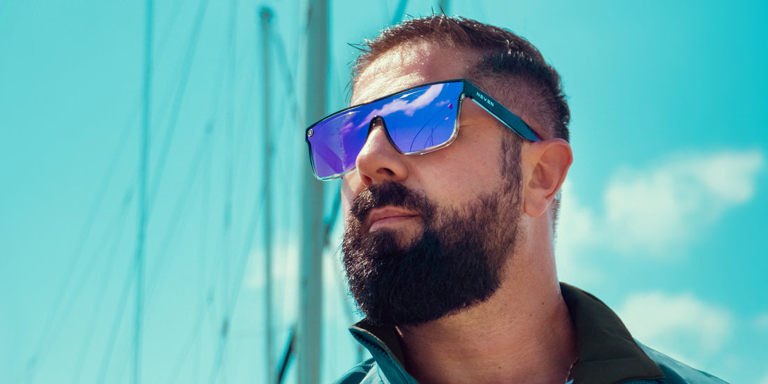 Man wearing Nomads XL sunglasses looking into the distance, close-up profile showing blue-purple mirrored lenses and sailboat masts against a teal sky