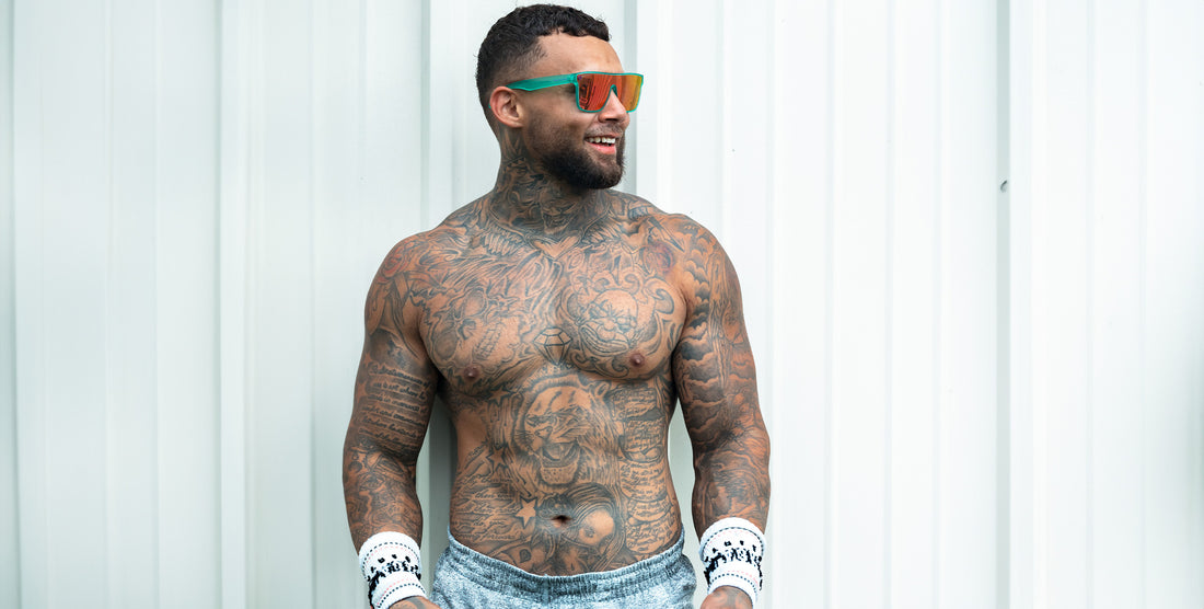 Tattooed shirtless man wearing Legend X teal-frame sunglasses with pink polarized lens, smiling against a white corrugated wall