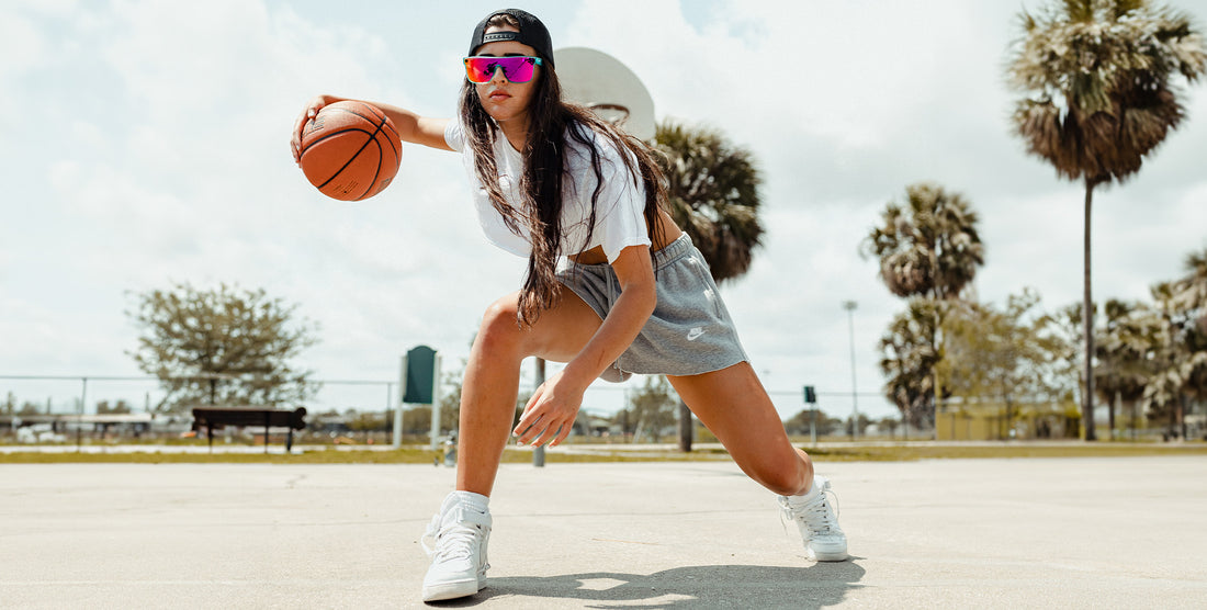 Young woman dribbling a basketball on an outdoor court, wearing transparent teal-frame X sunglasses with pink polarized lenses