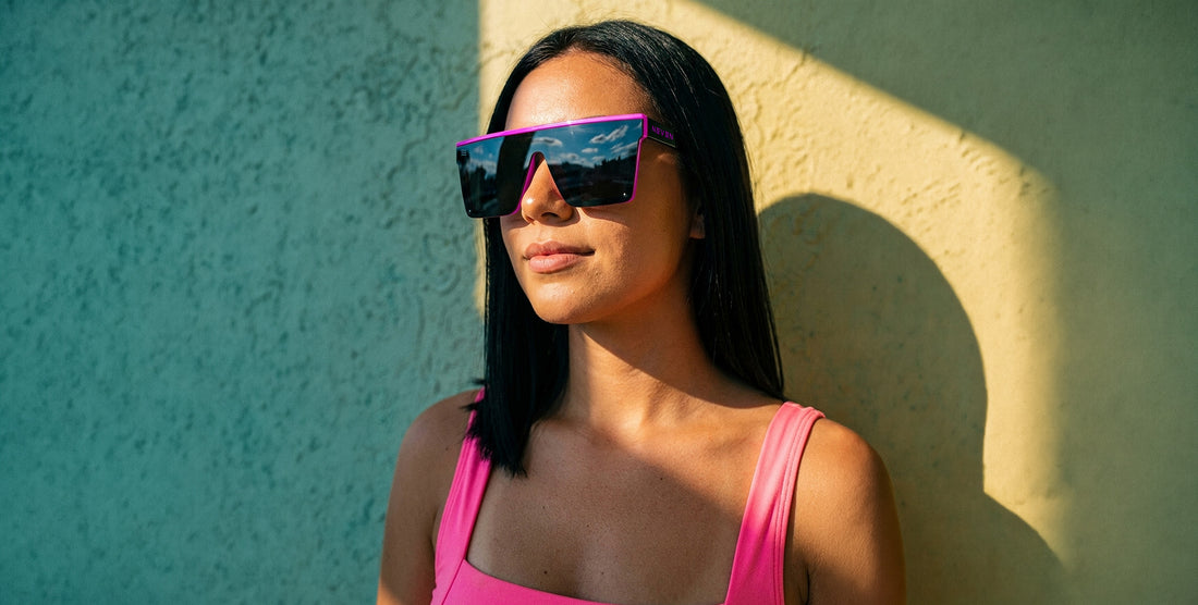 Woman wearing oversized pink-framed sunglasses against a sunlit textured wall, sky reflected in the lenses