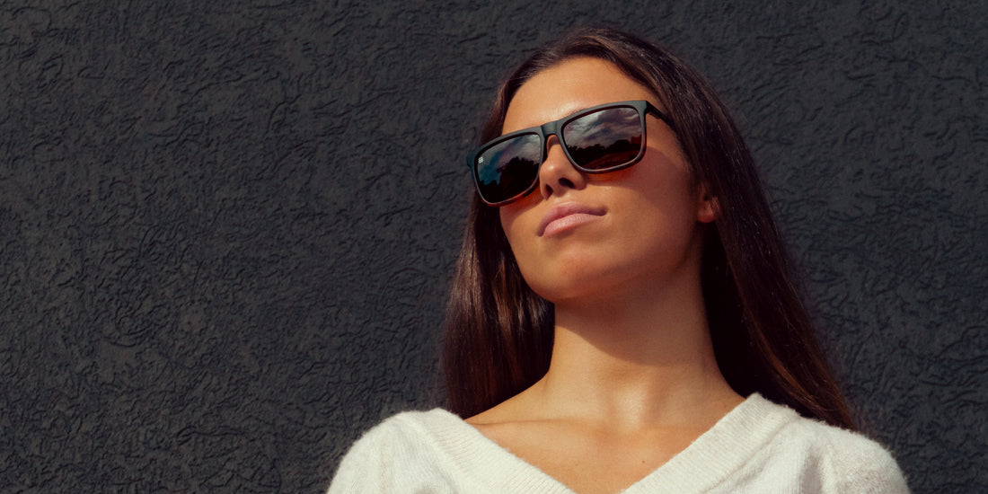 Woman wearing Repeater sunglasses with black frame, tortoiseshell splatter and brown lenses, looking up against a textured dark wall